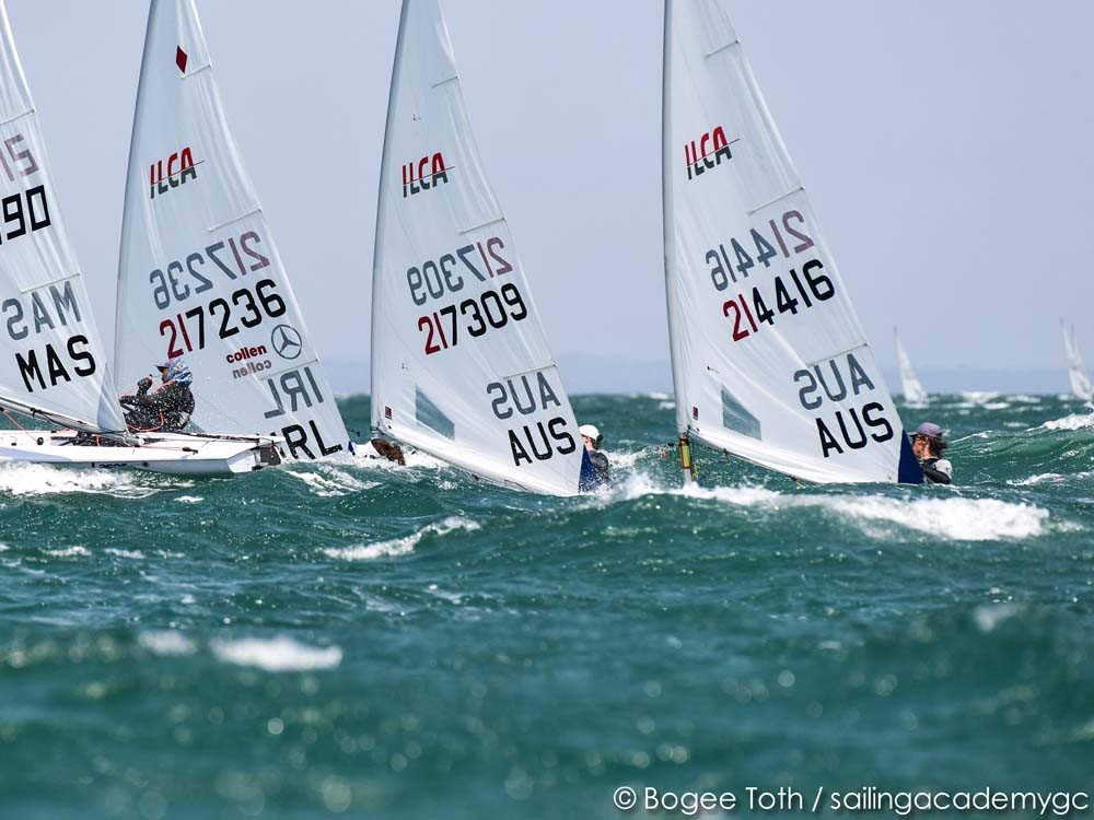 Régate dériveurs ILCA naviguant sur des vagues turquoise, voiles blanches avec inscriptions et logos, vue horizon légère.